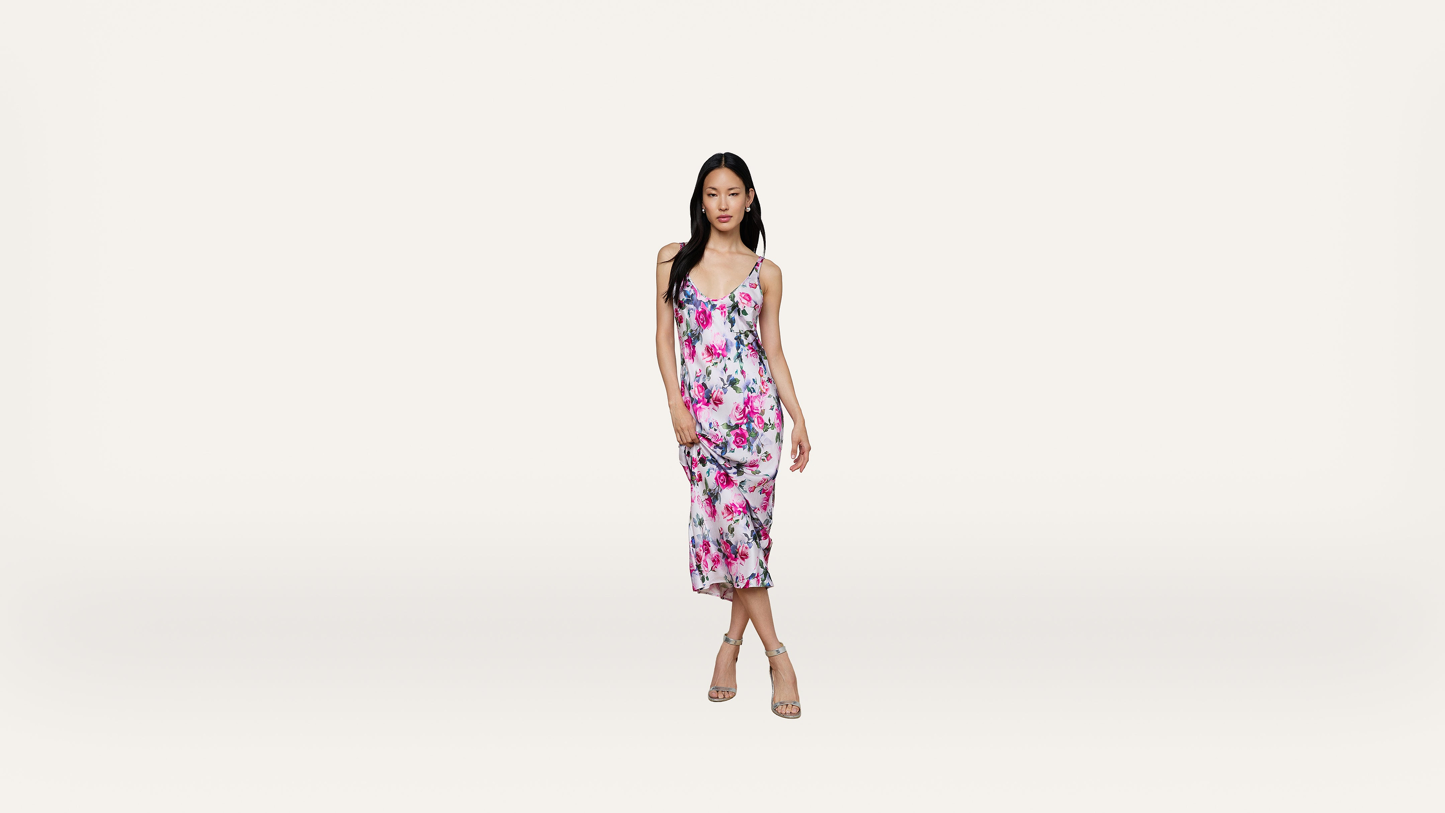 A woman wearing a sleeveless, floral-print dress and heels stands against a plain white background.