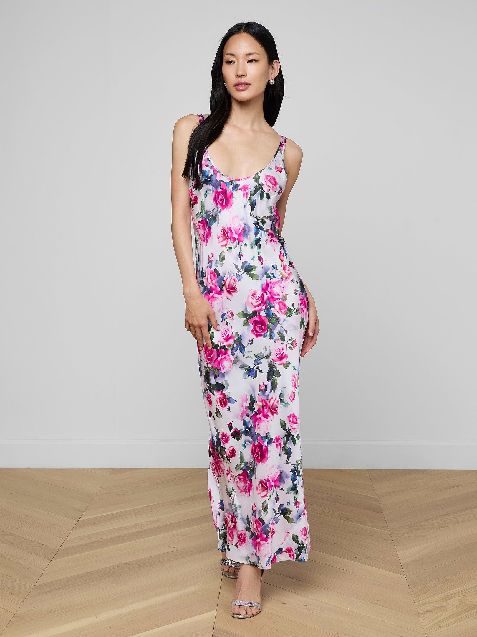 A woman wearing the Akiya Satin Maxi Dress—a sleeveless, white, floor-length bias-cut gown adorned with a pink and blue floral pattern—stands against a plain off-white background.