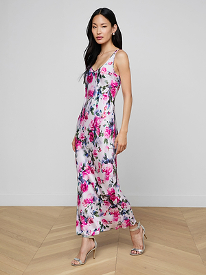 A woman stands on a wooden floor wearing the Akiya Satin Maxi Dress—a sleeveless, ankle-length bias-cut style in vibrant pink rose print—paired with silver high-heeled sandals. She faces slightly sideways against a plain light backdrop.