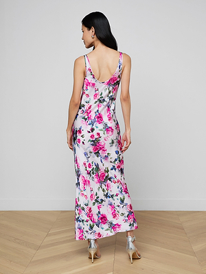 A woman with long dark hair stands on a wooden floor, facing away, wearing the sleeveless Akiya Satin Maxi Dress with a pink rose print and silver high-heeled sandals.