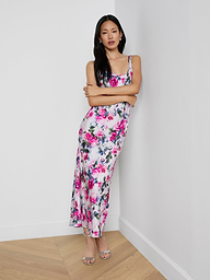 A woman stands against a white wall with arms crossed, wearing the Akiya Satin Maxi Dress in a pink rose print with magenta and white flowers, paired with silver open-toe heels on a light wood floor.