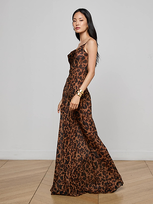 A woman in the Christine Silk Dress by L'AGENCE, a long sleeveless leopard print gown, stands sideways on a wooden floor against a white wall. She has straight dark hair and gold bracelets—an effortlessly chic evening look.