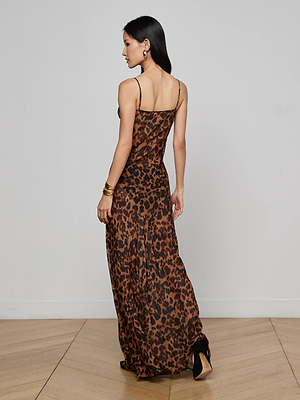 A woman with long dark hair stands on a wooden floor, wearing the L'AGENCE Christine Silk Dress—a long leopard print gown with thin straps—paired with black heels, gold earrings, and bracelets for an effortlessly chic evening look.