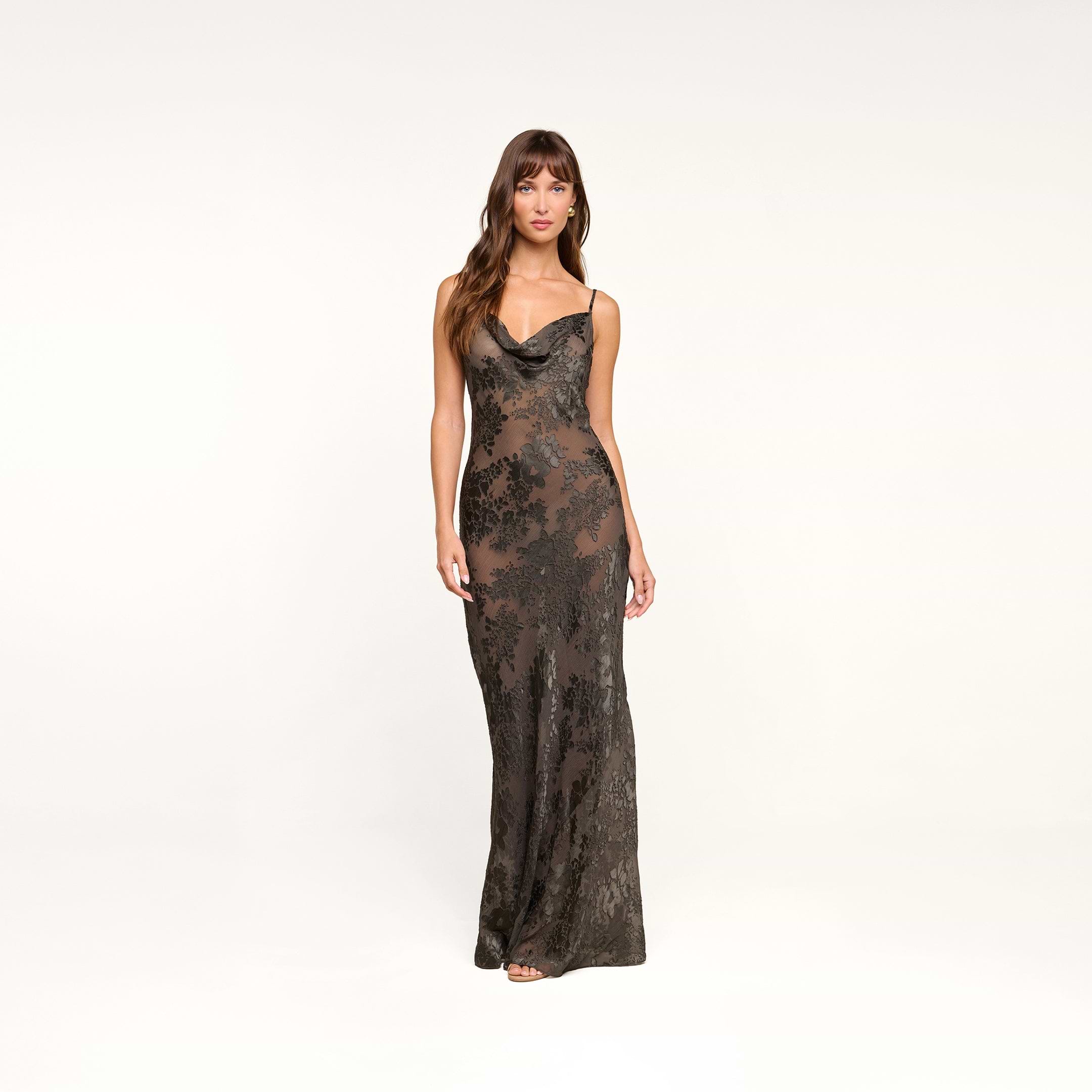 A woman stands against a plain background wearing the Christine Burnout Satin Gown—a long, fitted black dress with thin straps and a subtle floral pattern, reminiscent of a sheer slip dress.