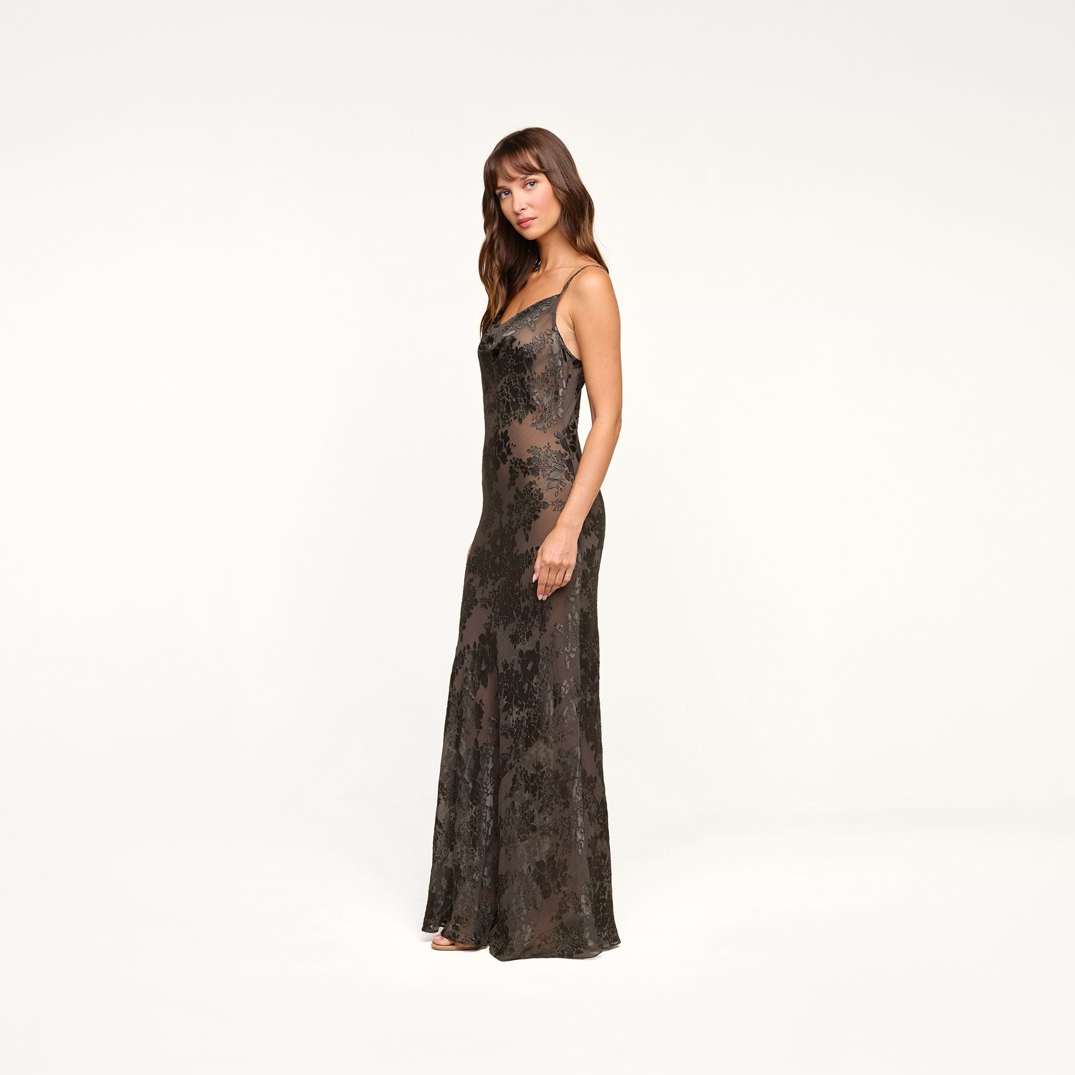 A woman in the Christine Burnout Satin Gown stands against a plain white background, facing slightly to the side with a neutral expression. Her elegant look evokes the essence of a sheer slip dress style.