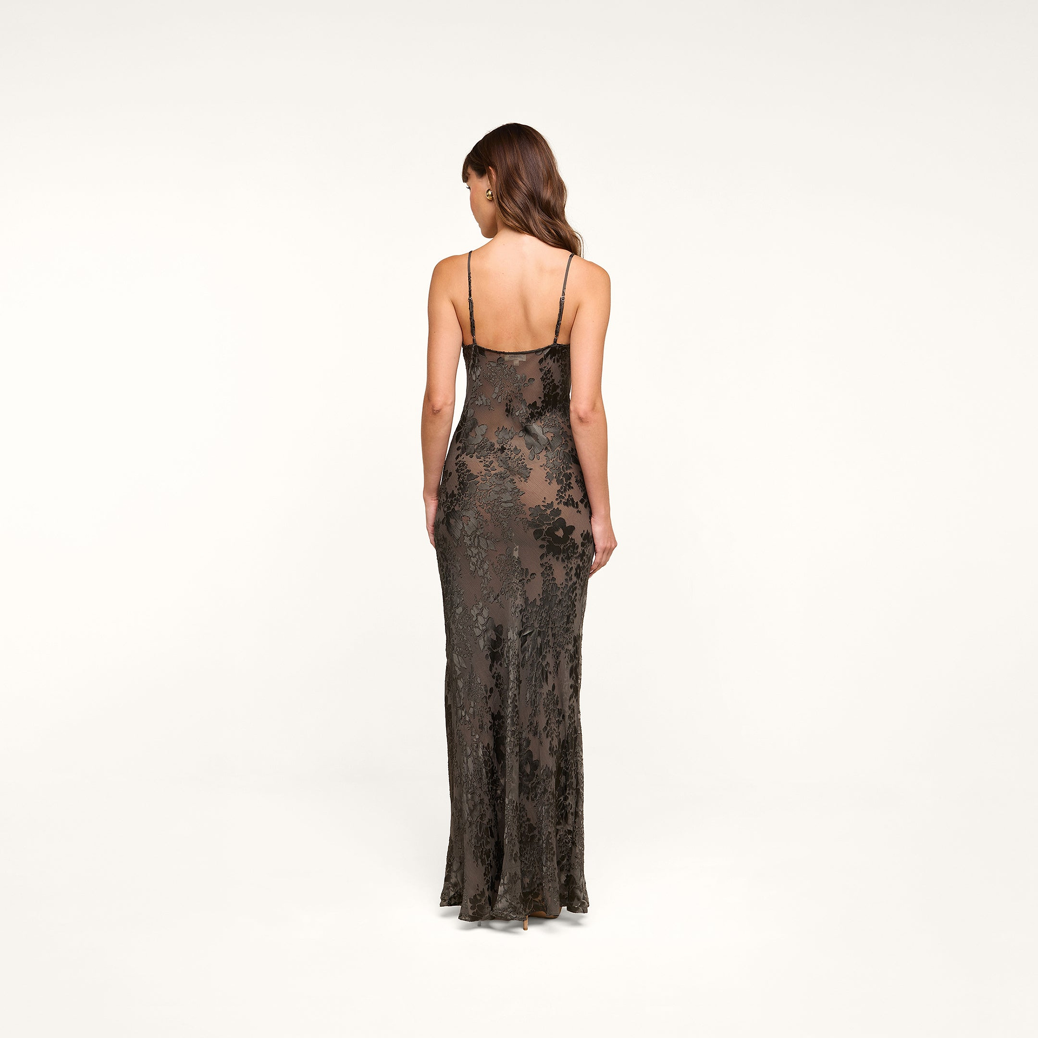 A woman stands facing away, wearing the Christine Burnout Satin Gown with its thin straps. Her hair is styled in loose waves, and the background is plain white.