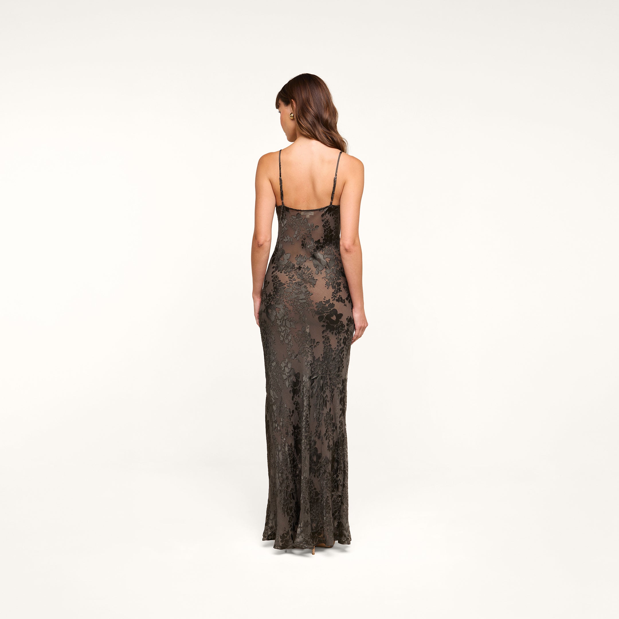 A woman stands facing away, wearing the Christine Burnout Satin Gown with its thin straps. Her hair is styled in loose waves, and the background is plain white.
