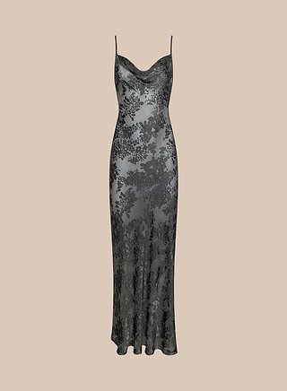 The Christine Burnout Satin Gown is a long, sleeveless slip dress with thin straps and a subtle cowl neckline, featuring delicate black floral lace over light gray fabric. Elegantly bias-cut, this sheer gown is displayed on a beige background.