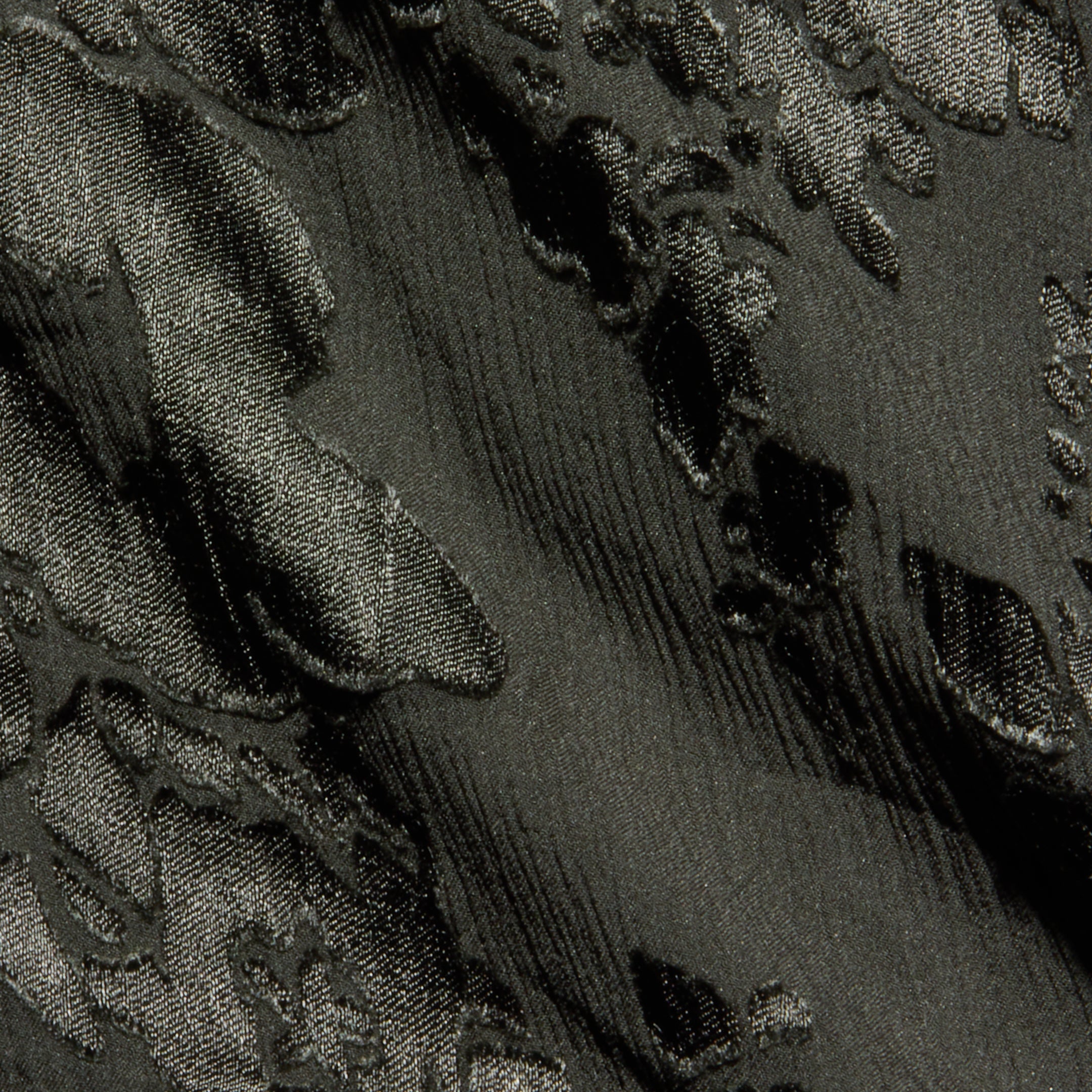 Close-up of textured dark fabric with raised, shiny floral or leaf patterns and a subtle sheen. The material features an embossed or jacquard weave design, similar to those found in the Christine Burnout Satin Gown.