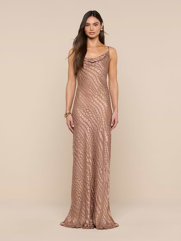 A woman stands against a beige background wearing the Christine Burnout Satin Gown, a long, sleeveless dress featuring a subtle textured pattern and a cowl neckline. She has long, straight brown hair and minimal accessories.