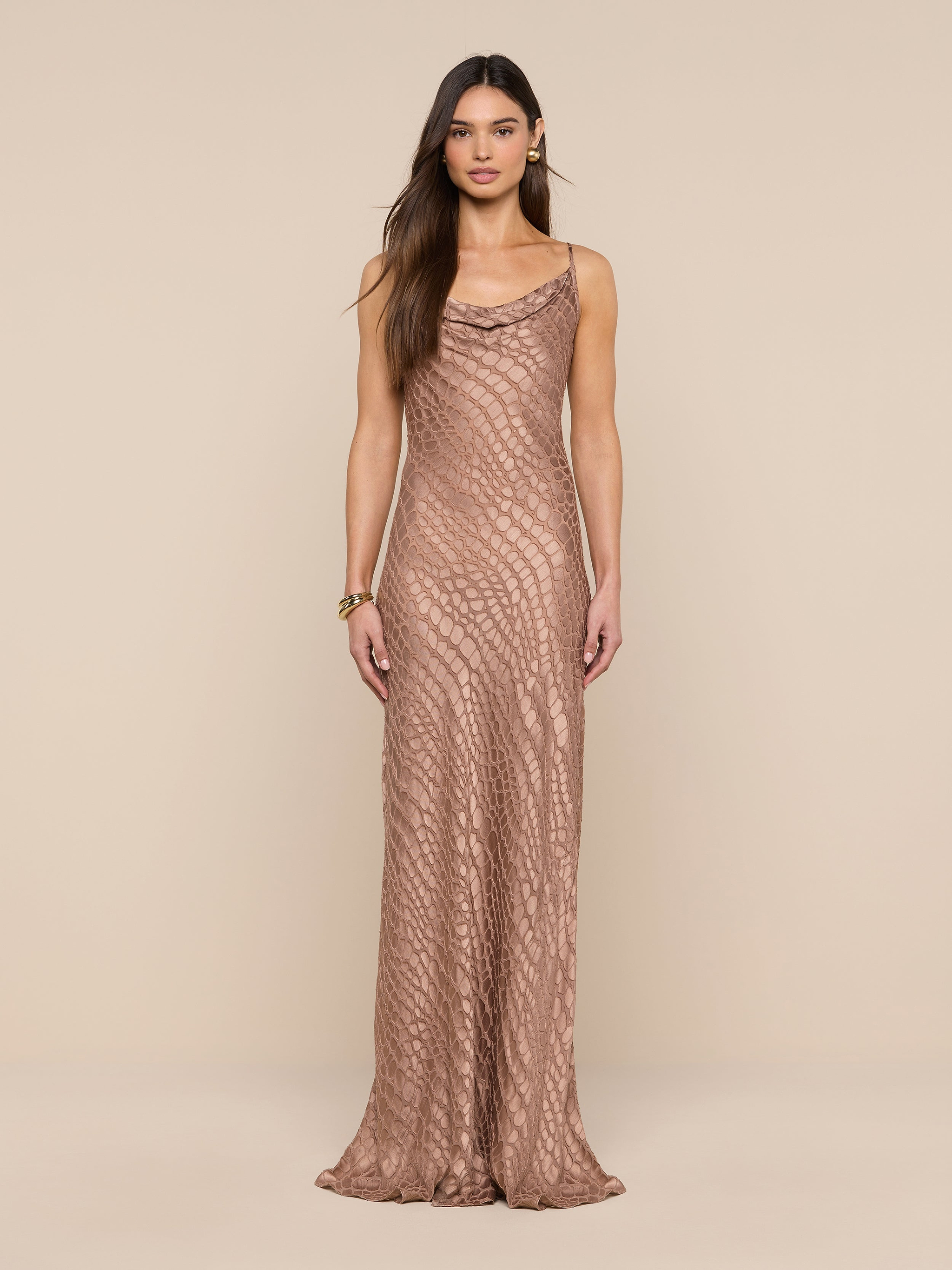 A woman stands against a beige background wearing the Christine Burnout Satin Gown, a long, sleeveless dress featuring a subtle textured pattern and a cowl neckline. She has long, straight brown hair and minimal accessories.