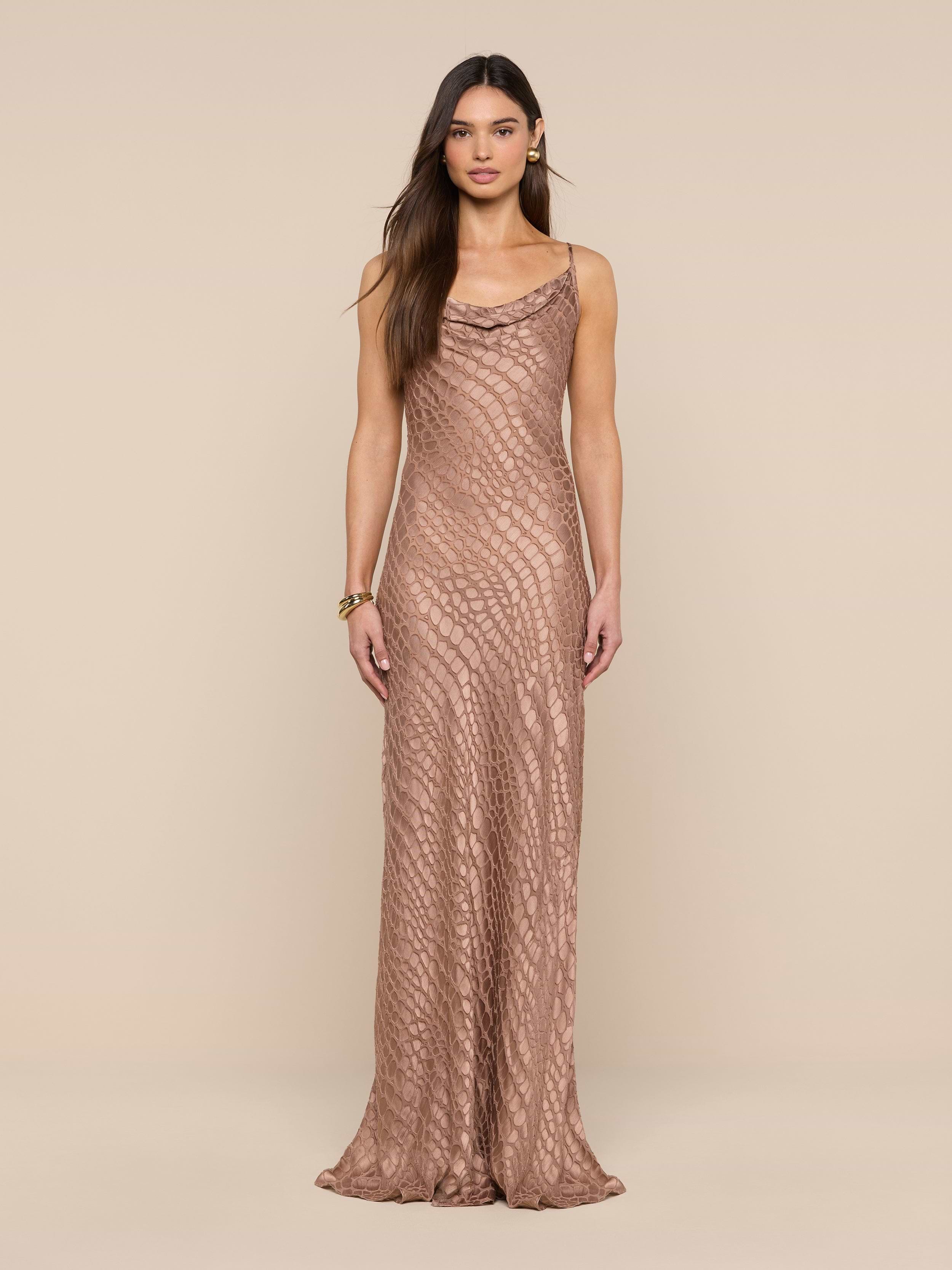 A woman stands against a beige background wearing the Christine Burnout Satin Gown, a long, sleeveless dress featuring a subtle textured pattern and a cowl neckline. She has long, straight brown hair and minimal accessories.