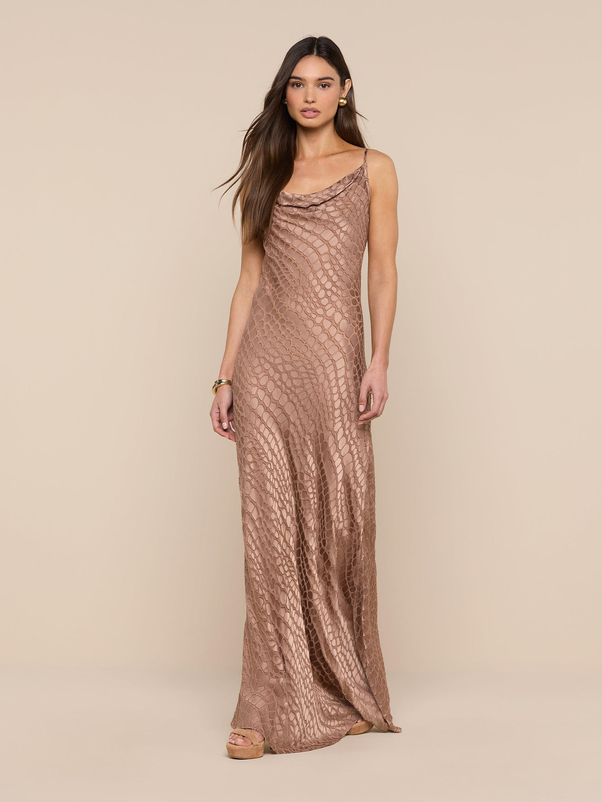 A woman wearing the Christine Burnout Satin Gown—a sleeveless, shimmery bronze dress with a subtle scale-like pattern—stands against a plain beige background.