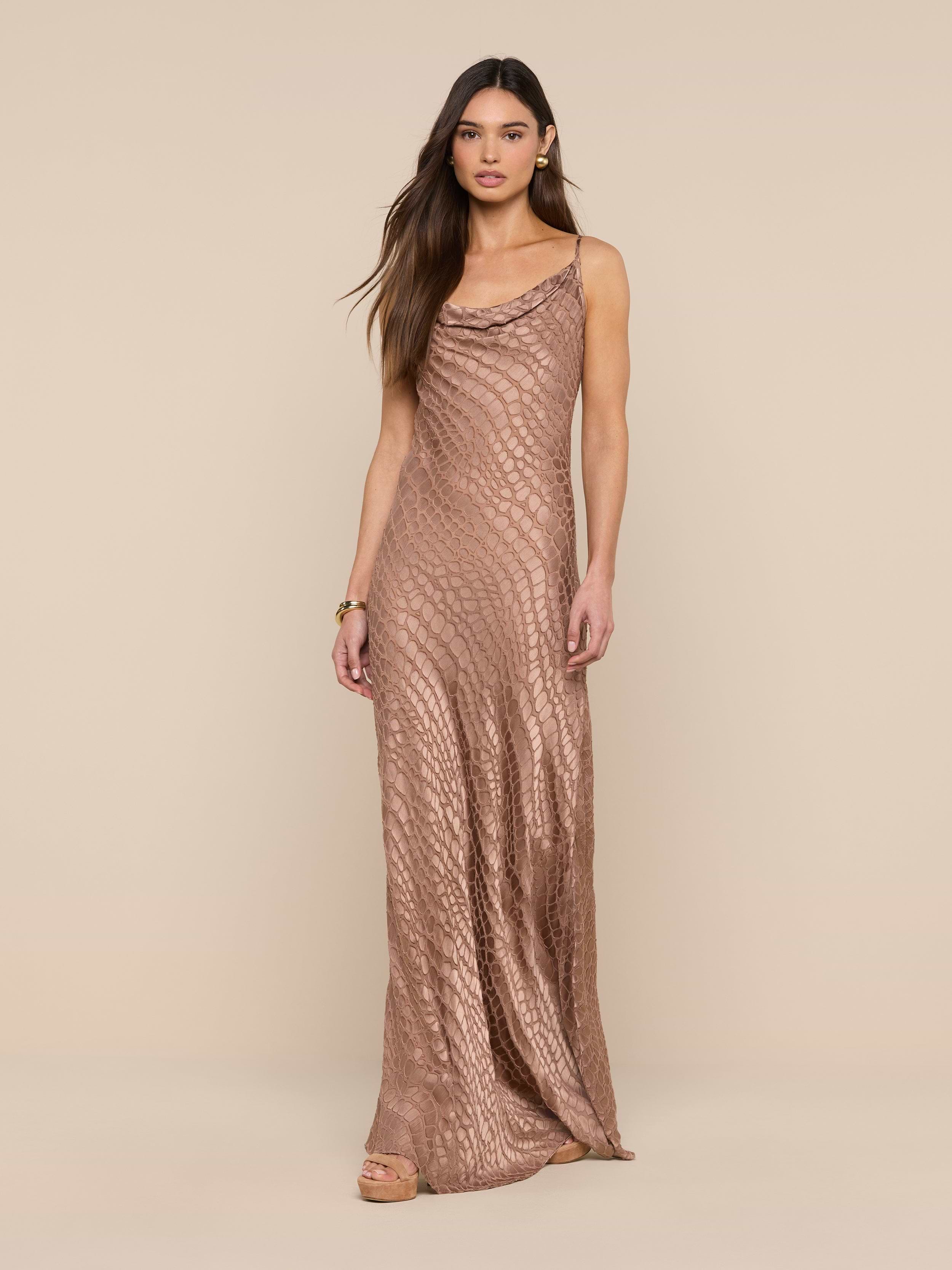 A woman wearing the Christine Burnout Satin Gown—a sleeveless, shimmery bronze dress with a subtle scale-like pattern—stands against a plain beige background.