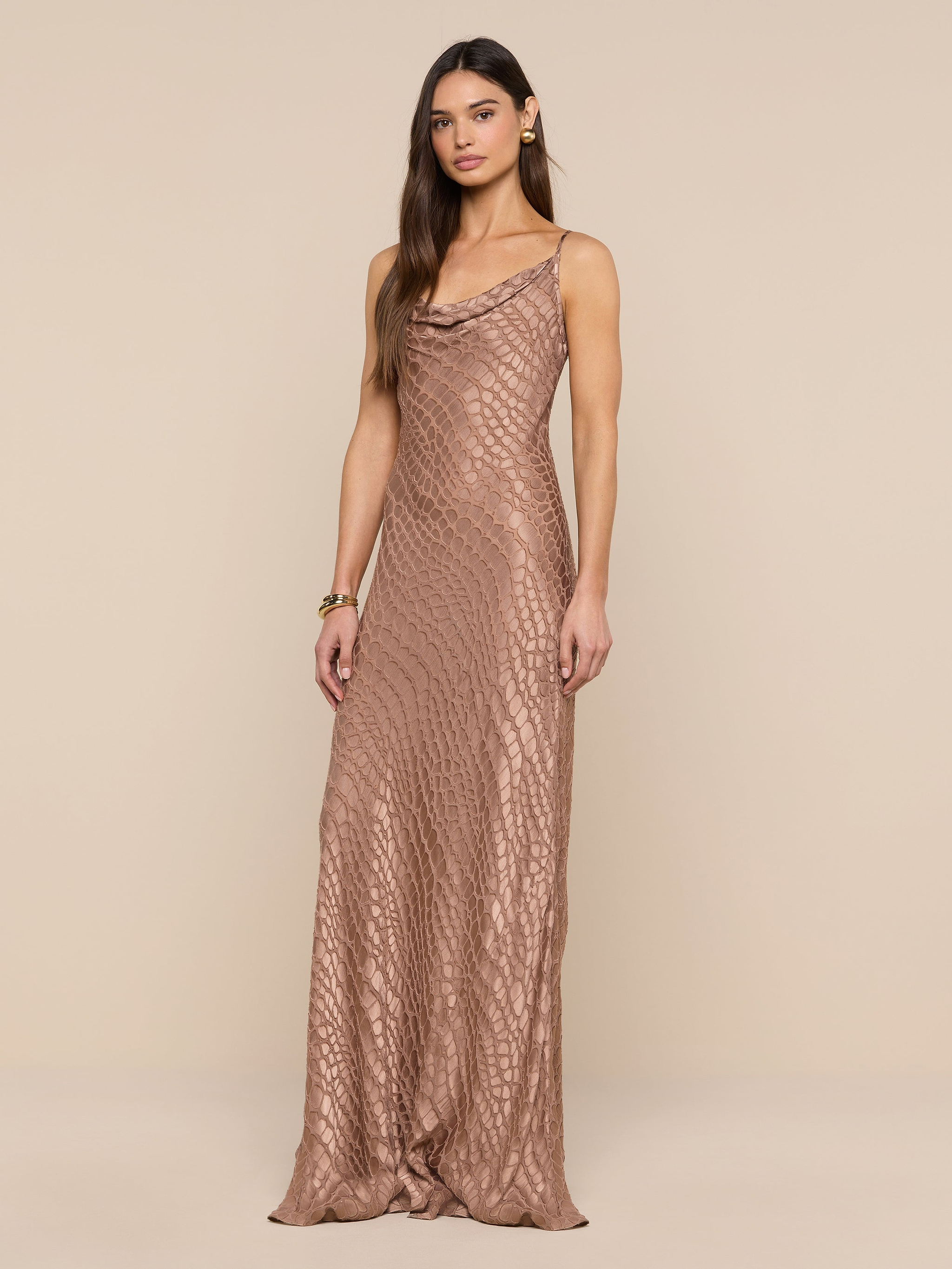 A woman stands against a beige background wearing the Christine Burnout Satin Gown, a sleeveless, floor-length dress crafted in shimmery bronze satin with a subtle scale-like pattern. She has long hair, minimal jewelry, and a neutral expression.