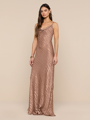 A woman with long brown hair wears the Christine Burnout Satin Dress, a sleeveless, floor-length metallic bronze gown with a subtle scale-like pattern, standing against a beige background.