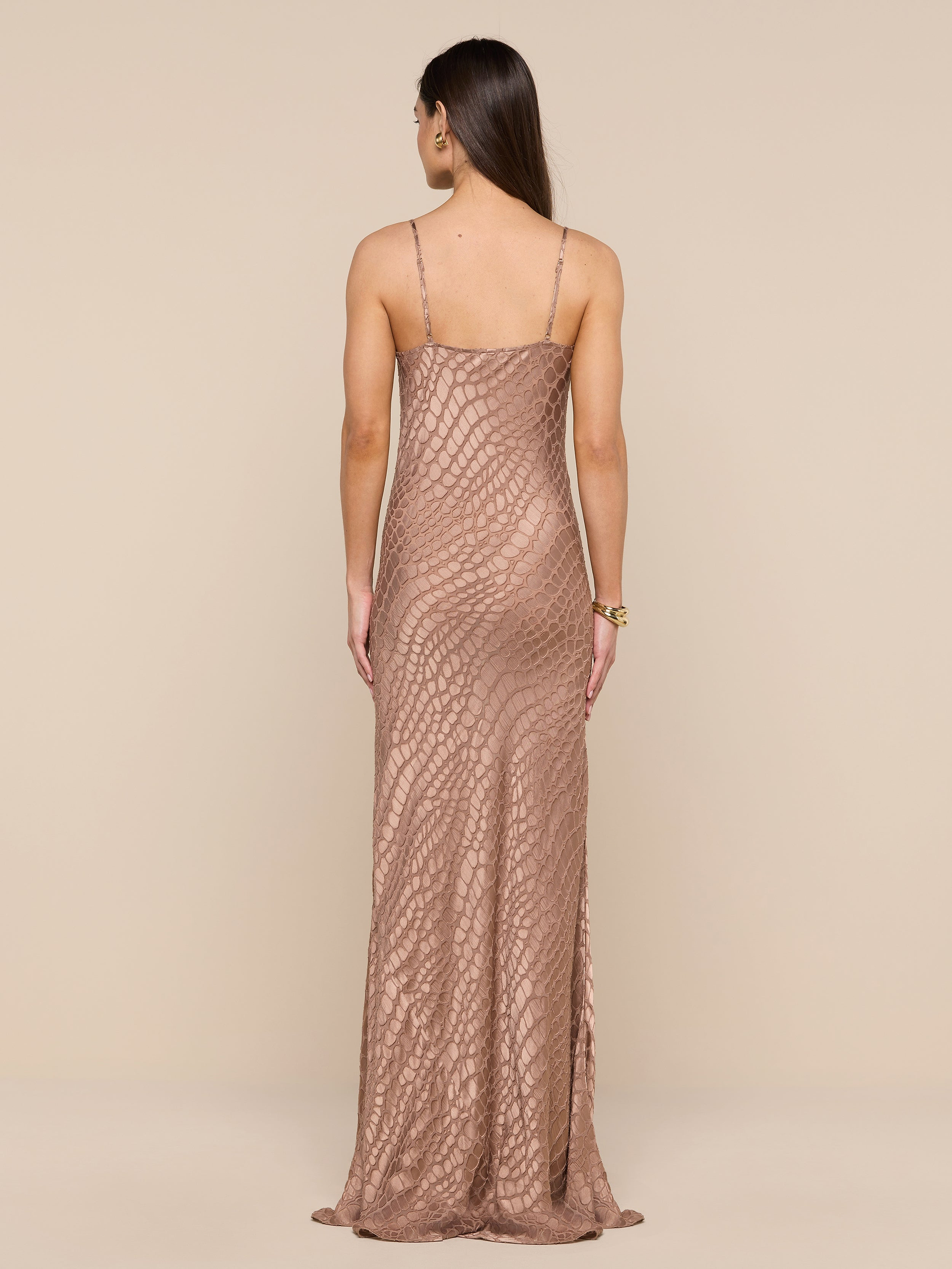 A woman with long brown hair stands facing away against a neutral background, wearing the Christine Burnout Satin Gown—a long, fitted, sleeveless dress in rose-gold featuring a textured geometric pattern.