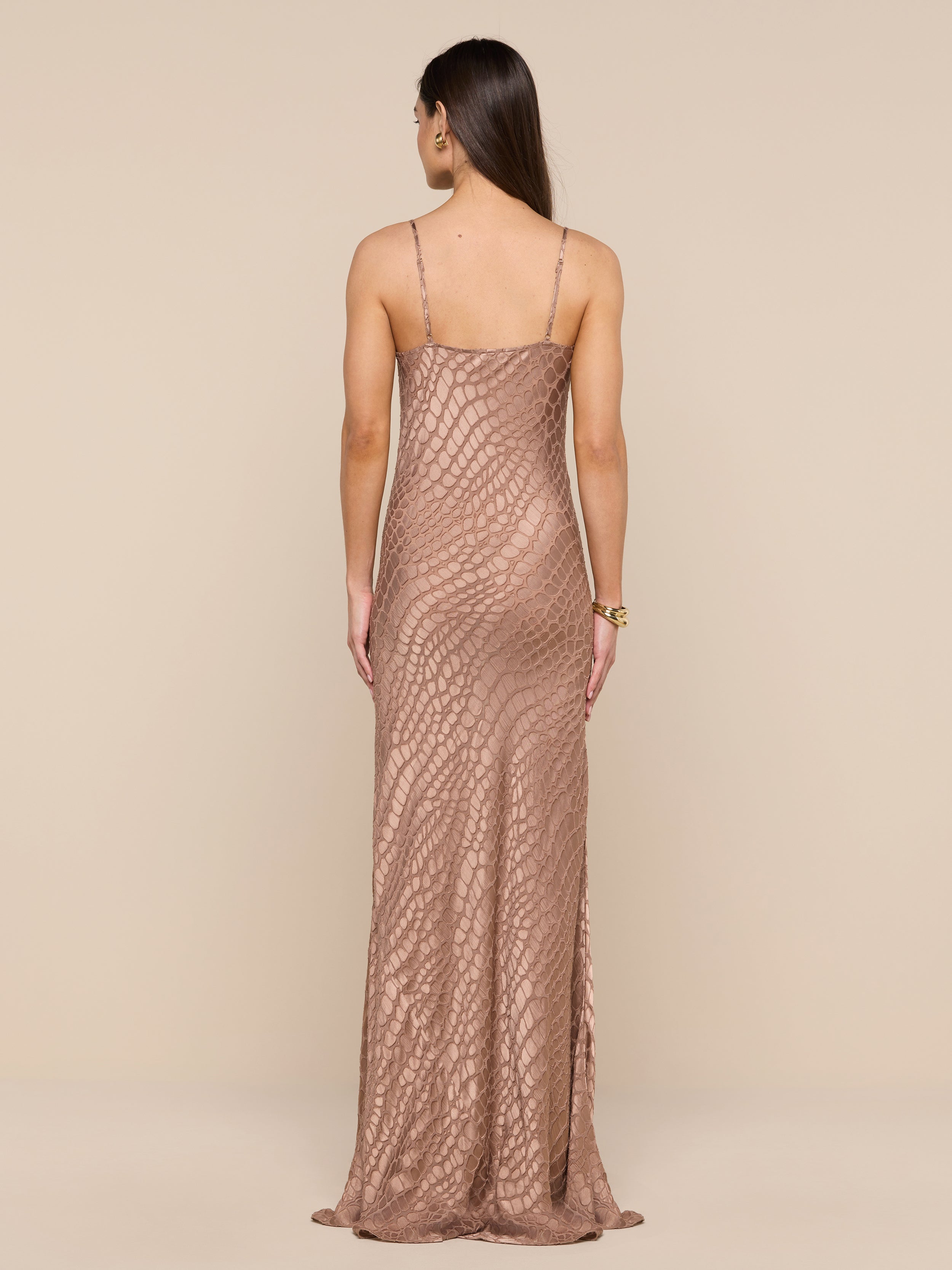 A woman with long brown hair stands facing away against a neutral background, wearing the Christine Burnout Satin Gown—a long, fitted, sleeveless dress in rose-gold featuring a textured geometric pattern.