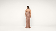 A woman with long brown hair, wearing a shimmering, floor-length, backless gown, stands facing away against a plain white background.