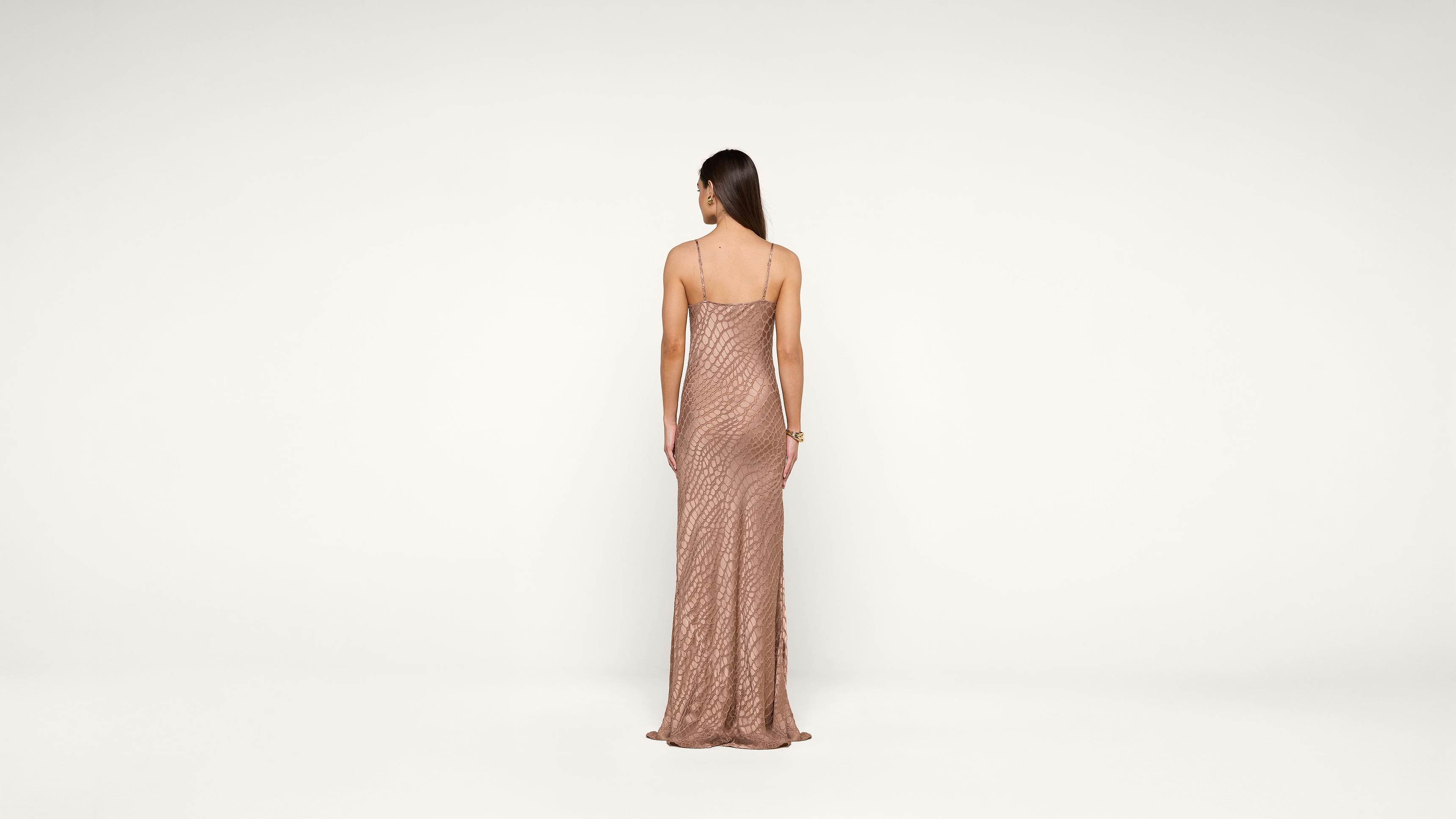 A woman with long brown hair, wearing a shimmering, floor-length, backless gown, stands facing away against a plain white background.
