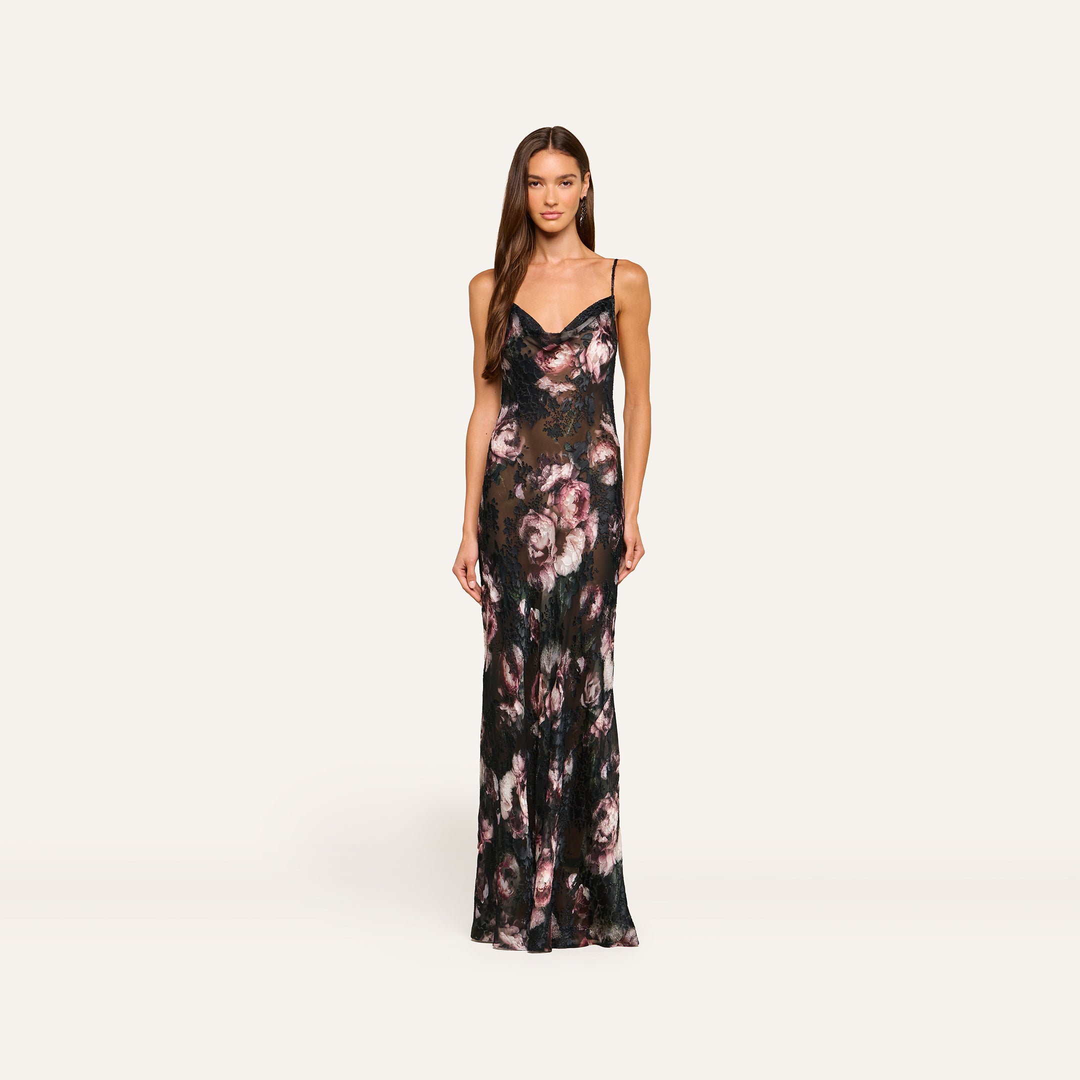 A woman wearing the Christine Dress—a long, fitted floral dress with thin straps and dark, muted pink rose patterns—stands against a plain light background.
