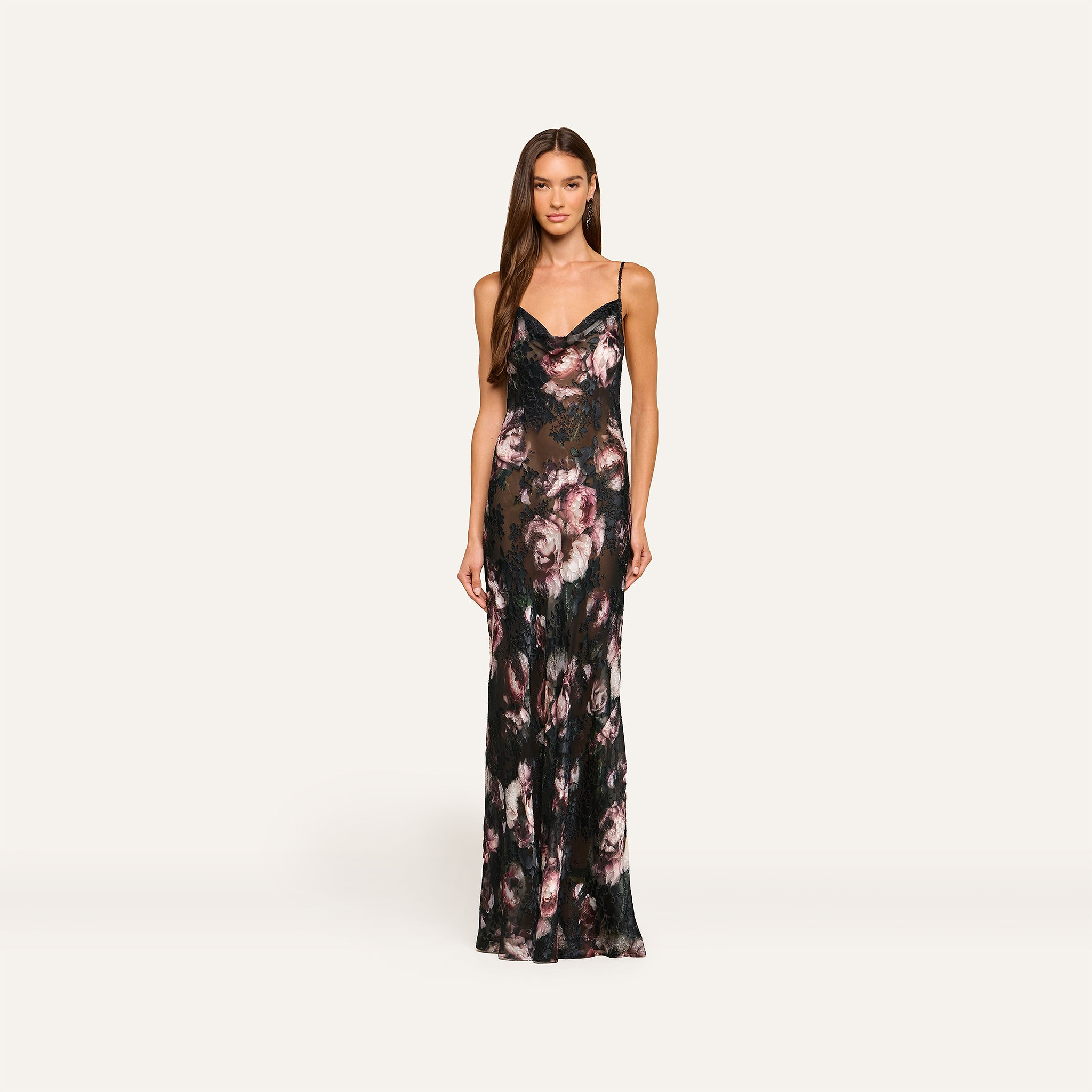 A woman wearing the Christine Dress—a long, fitted floral dress with thin straps and dark, muted pink rose patterns—stands against a plain light background.