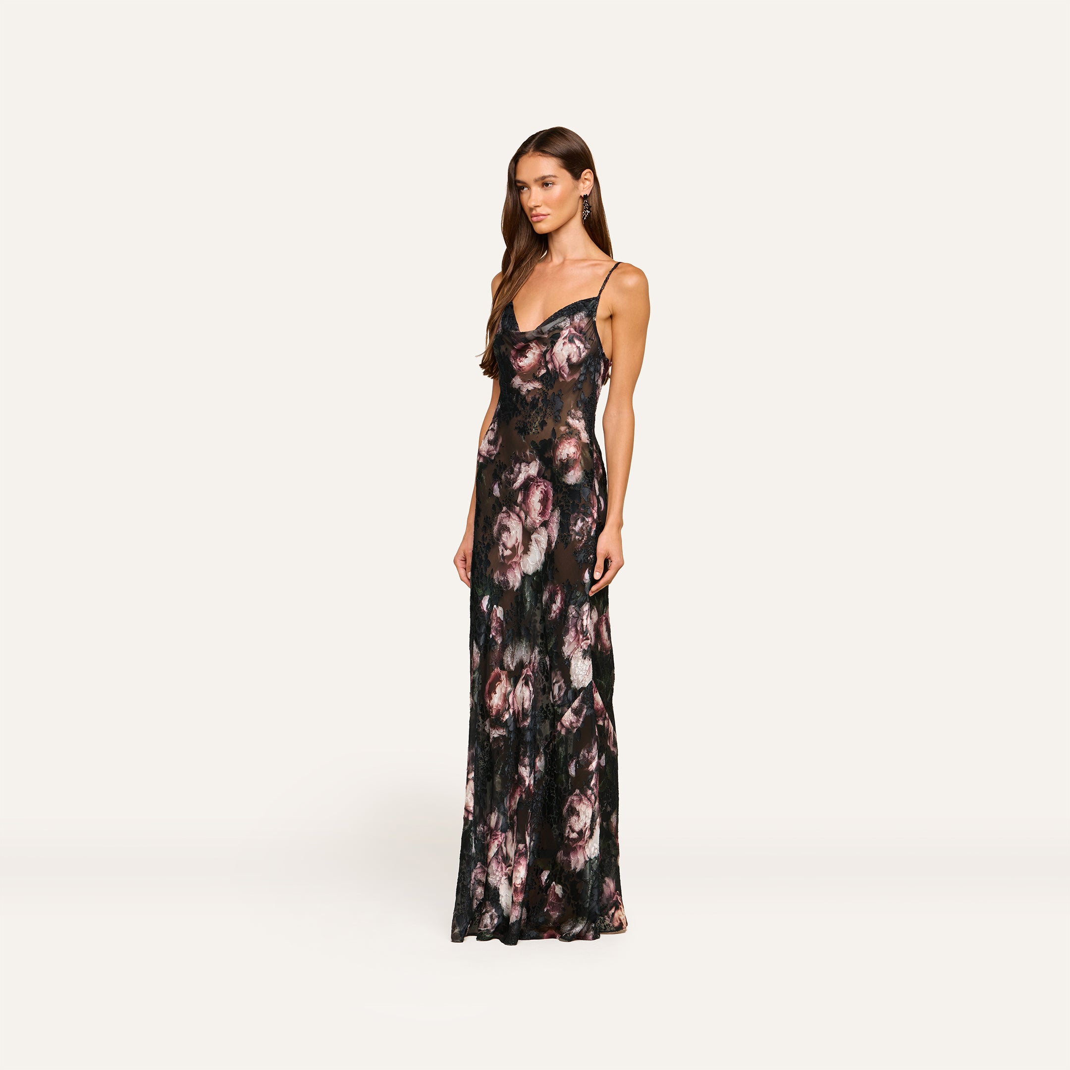 A woman stands against a plain light background, wearing the Christine Dress—a long, sleeveless black burnout satin dress adorned with a pink and purple rose print.