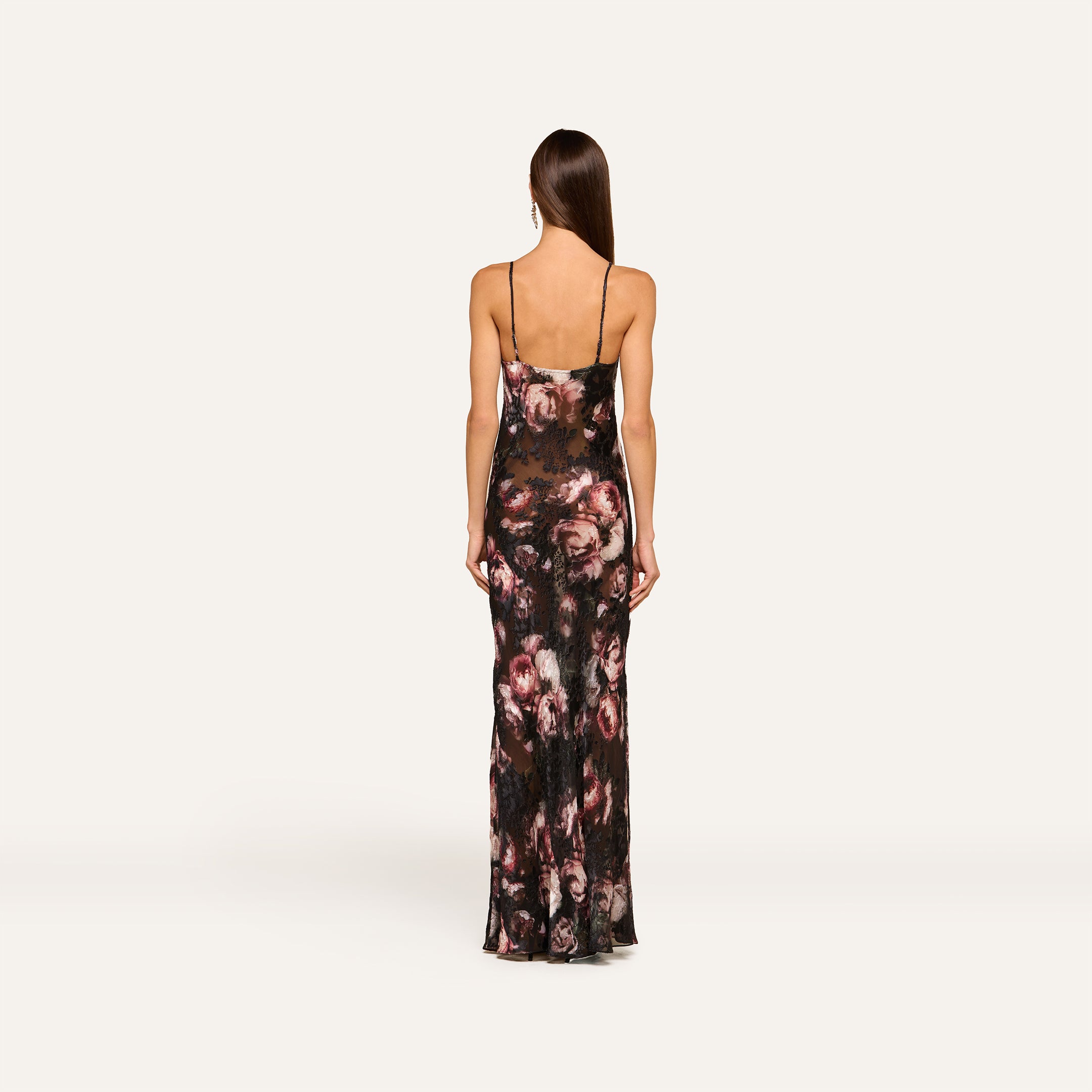 A woman stands facing away against a plain light background, wearing the Christine Dress—a long, fitted black dress with rose print, sleeveless design, and thin straps.