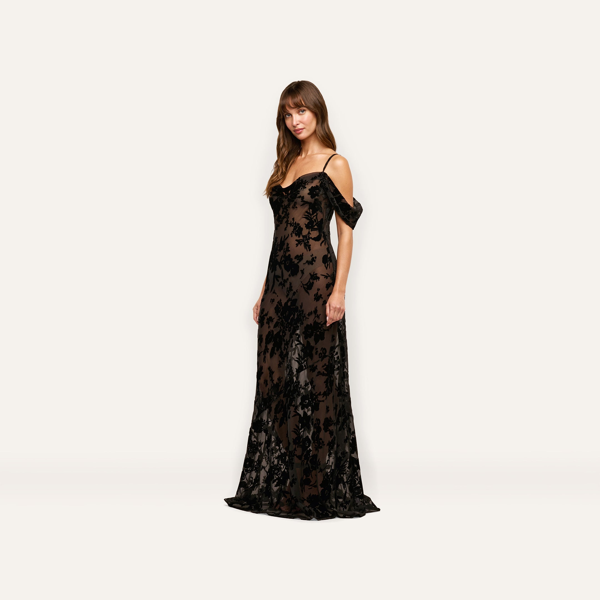 A woman wearing the Kenna Velvet Gown, a floor-length, sheer black burnout velvet dress with thin straps and off-the-shoulder sleeves.