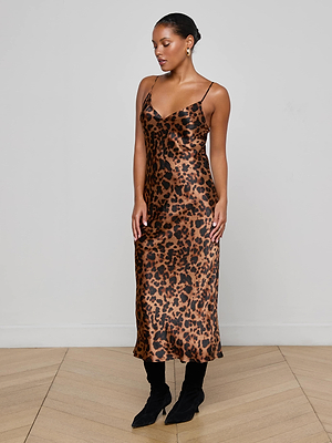 A woman poses indoors against a light wall and wooden floor, wearing the L'AGENCE Seridie Silk Slip Dress in a leopard print with thin straps, her hair pulled back, and styled with black boots.