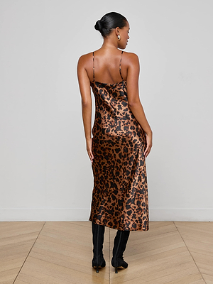A woman stands on a wooden floor, facing away. She wears the L'AGENCE Seridie Silk Slip Dress in leopard print with thin straps, black knee-high boots, and her hair styled in a neat bun. The background is plain and light-colored.