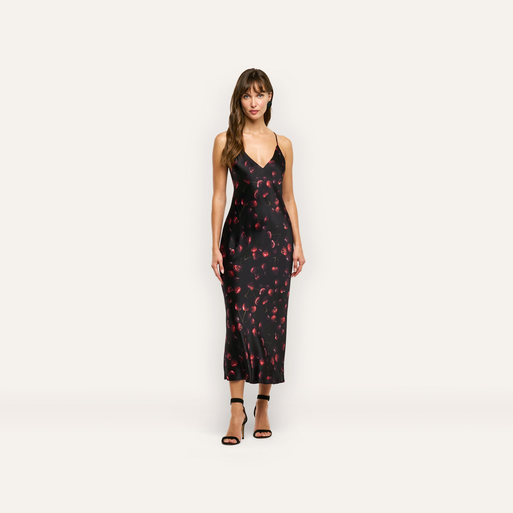 A woman wearing a black Seridie Silk Slip Dress in silk charmeuse with a red cherry print and black strappy high heels.