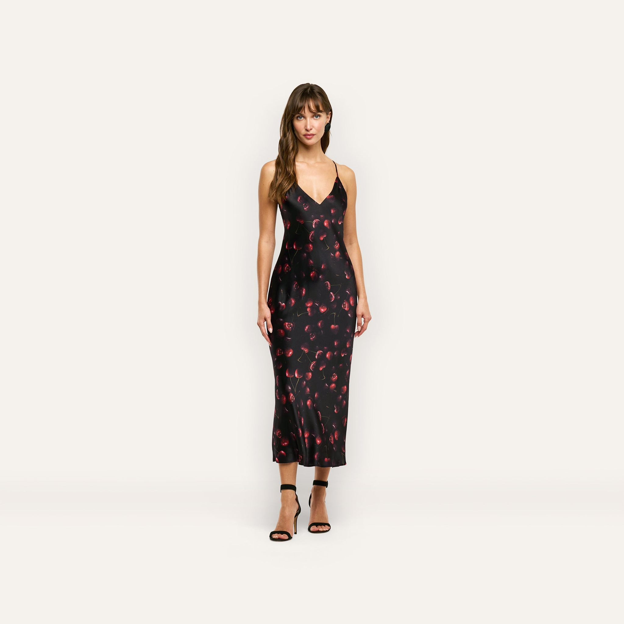 A woman wearing a black Seridie Silk Slip Dress in silk charmeuse with a red cherry print and black strappy high heels.