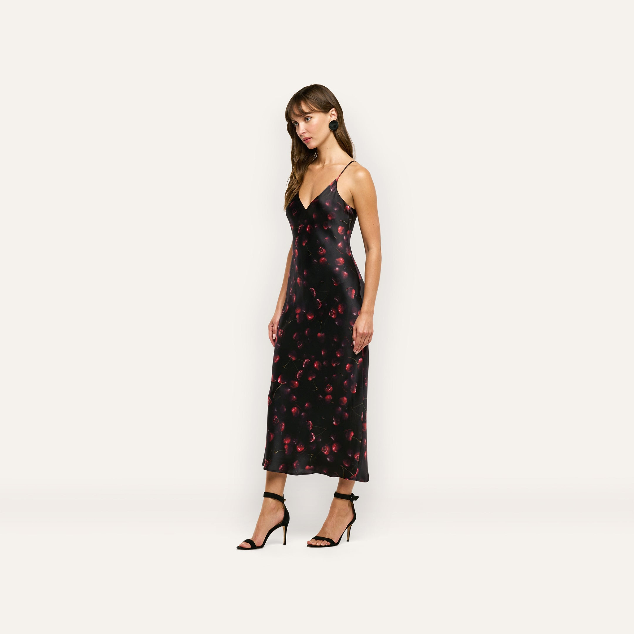 A woman wearing a black silk charmeuse Seridie Silk Slip Dress with a red cherry print and black heeled sandals.