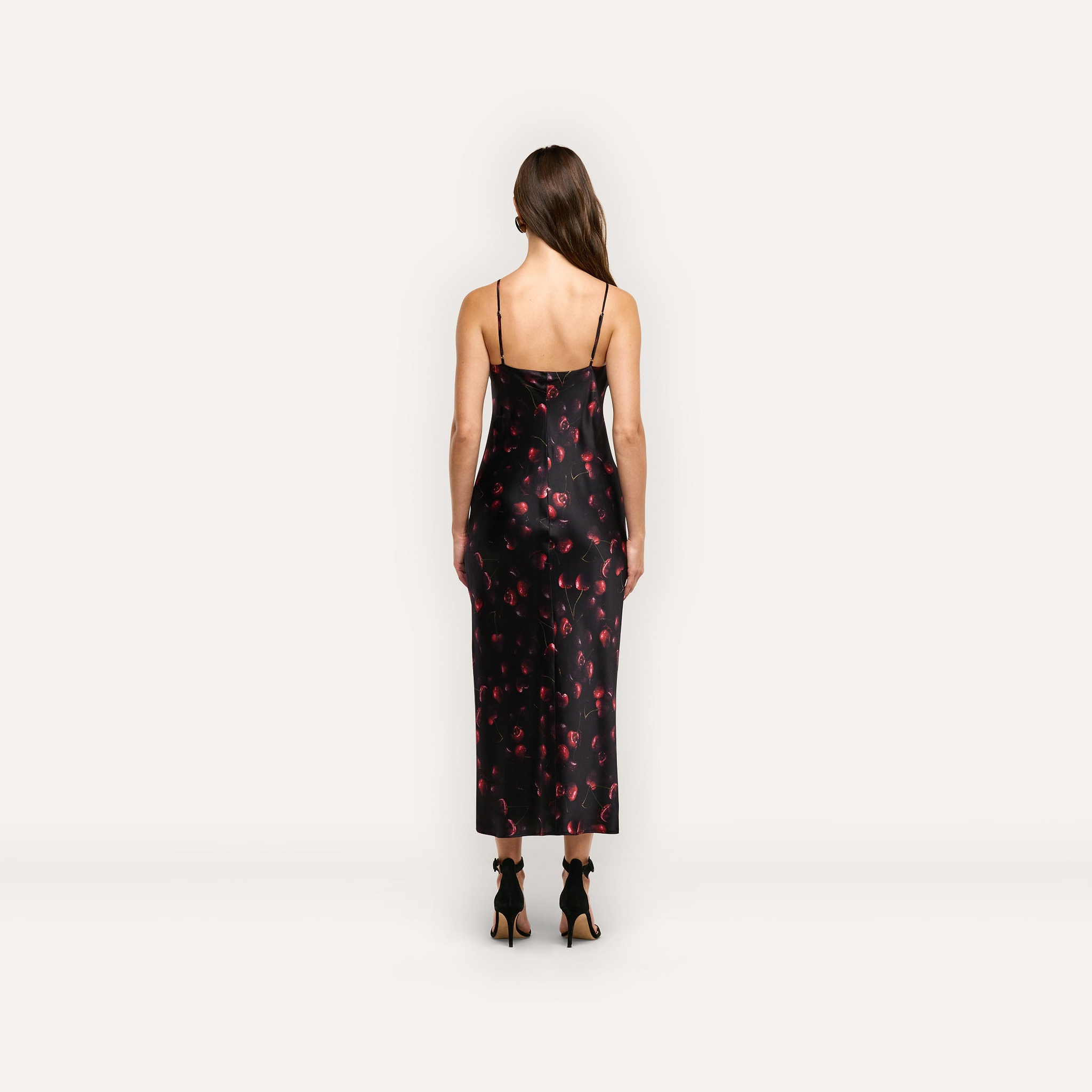Back view of a woman wearing a black cherry print Seridie Silk Slip Dress with thin spaghetti straps and black heeled sandals.