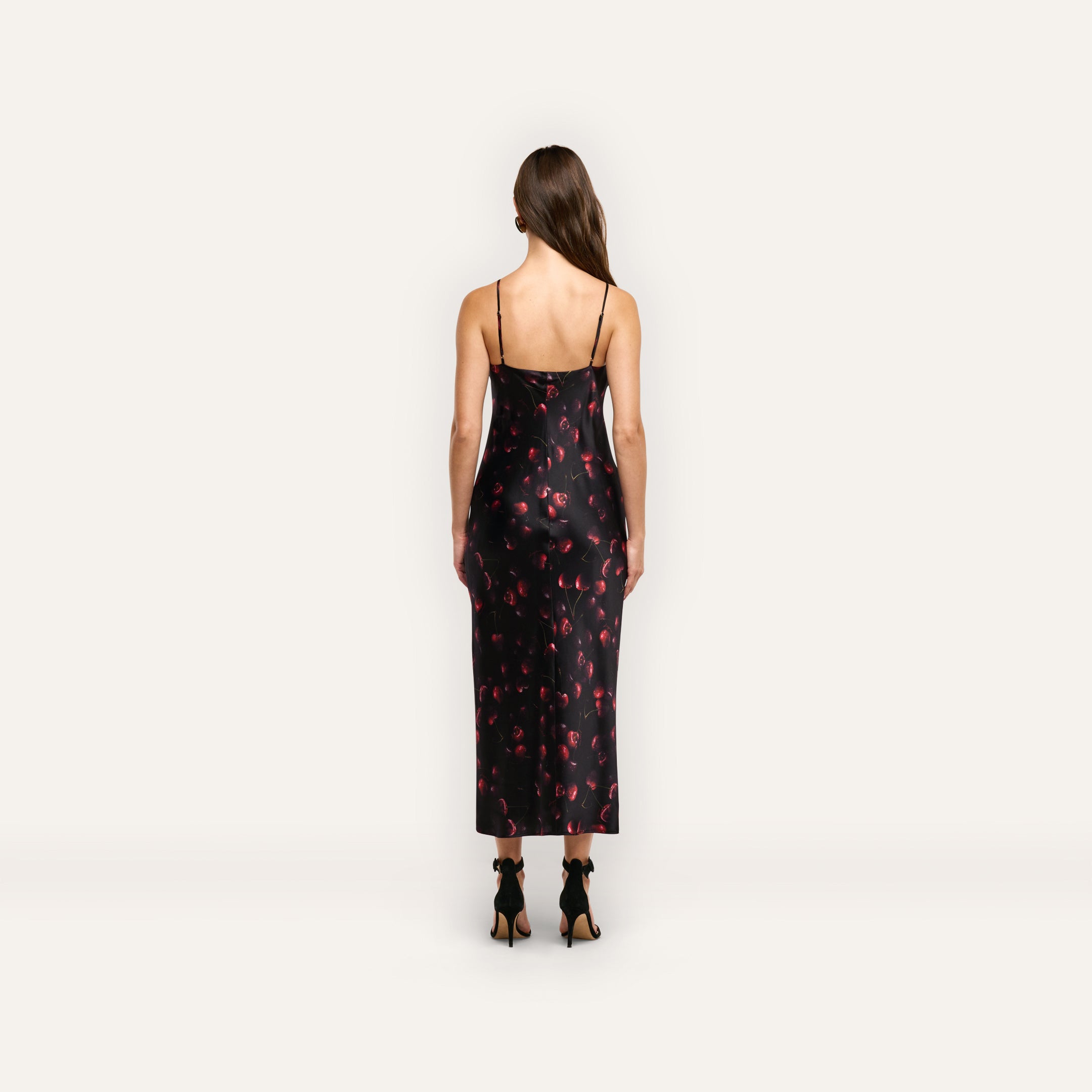 Back view of a woman wearing a black cherry print Seridie Silk Slip Dress with thin spaghetti straps and black heeled sandals.