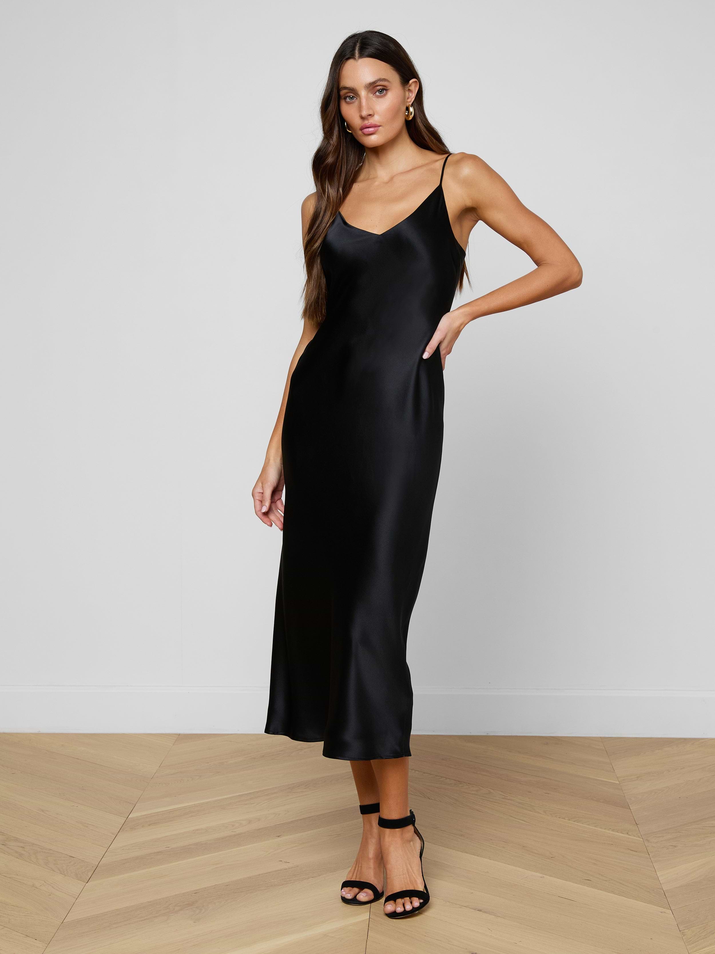 A woman with long, wavy brown hair and a relaxed pose models the Seridie Silk Slip Dress—black, sleeveless, with adjustable straps—paired with black ankle-strap heels. She stands on a wooden floor against a plain white wall.