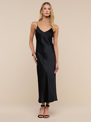 A woman in the Seridie Silk Slip Dress—a sleeveless, black silk design with thin straps—pairs it with black high-heeled sandals, gold earrings, and a bracelet. She stands against a beige background with long blonde hair.