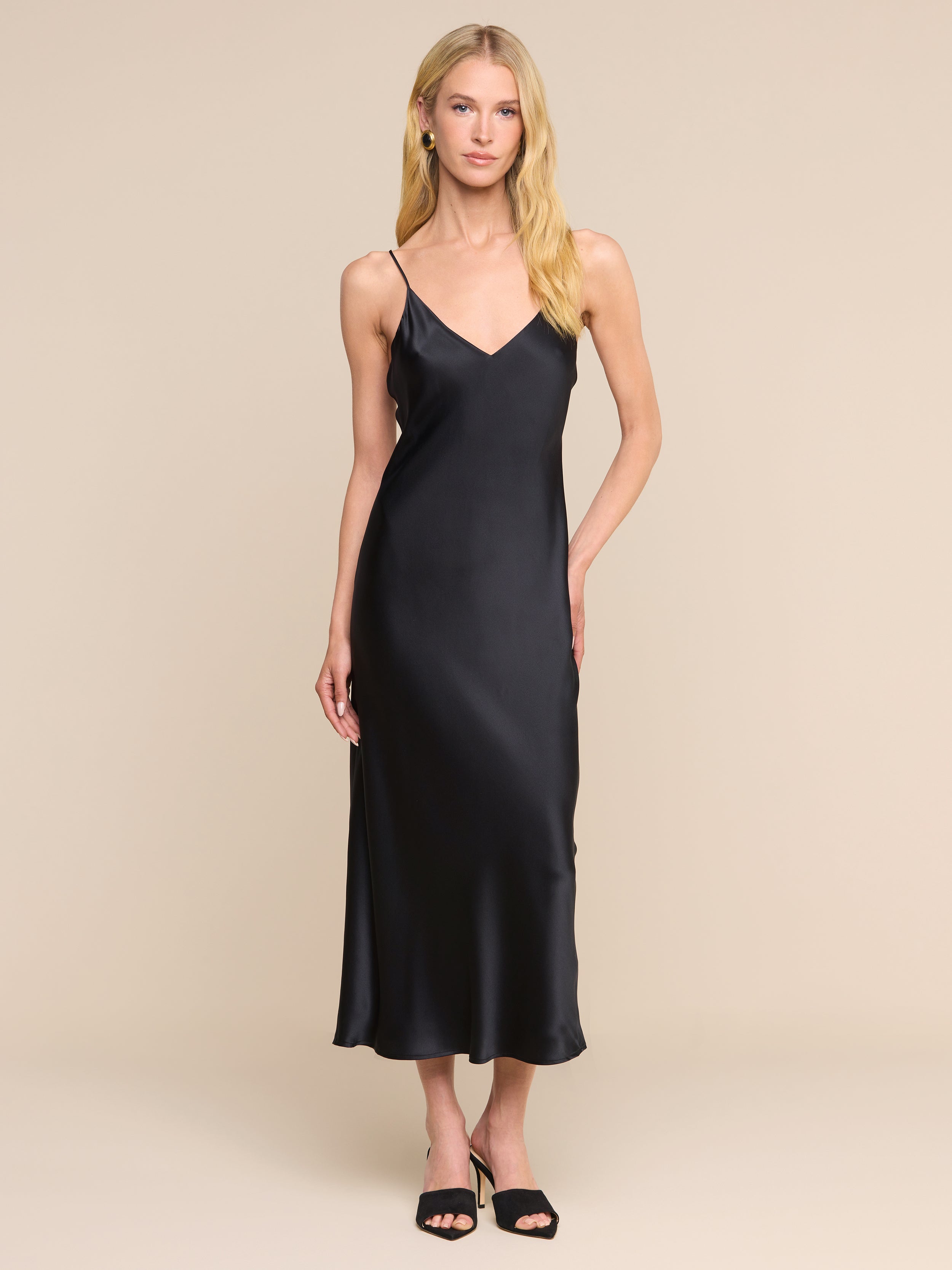 A woman with long blonde hair wears the Seridie Silk Slip Dress—a sleeveless, V-neck black design—paired with black heels, standing against a plain white background.