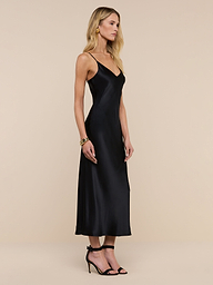A woman with long blonde hair wears the Seridie Silk Slip Dress, a black, sleeveless V-neck style, along with black heeled sandals, standing in profile against a plain white background.