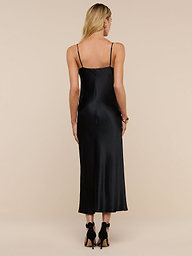 A person with long blonde hair wears the Seridie Silk Slip Dress—black, sleeveless, with thin straps—paired with black heels, standing against a plain white background and facing away from the camera.