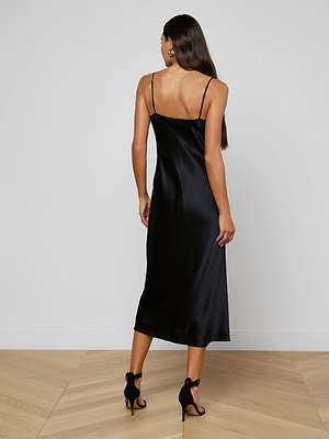 A woman with long brown hair stands on a wooden floor, facing away, wearing the Seridie Silk Slip Dress—black, mid-length, with adjustable straps—and black high-heeled sandals against a plain white background.