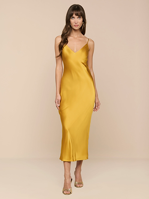 A woman wearing the Seridie Silk Slip Dress in gold with thin straps and strappy heels stands against a plain beige background.