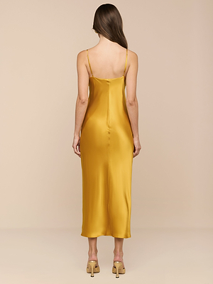 A woman with long brown hair stands facing away, wearing the Seridie Silk Slip Dress in gold with thin straps and beige heeled sandals, set against a plain beige background.