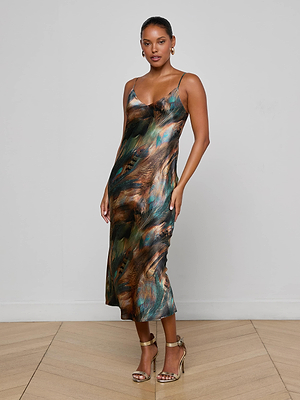 A woman poses on a wooden floor against a light gray wall, wearing the L’AGENCE Seridie Silk Slip Dress—bias-cut with an abstract pattern and thin straps—paired with gold high-heeled sandals.