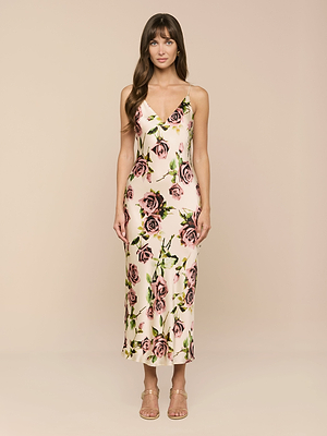 A woman with long brown hair and bangs stands against a beige background wearing the Seridie Silk Slip Dress—a sleeveless, light pink slip dress featuring a floral rose print—paired with strappy nude heels.