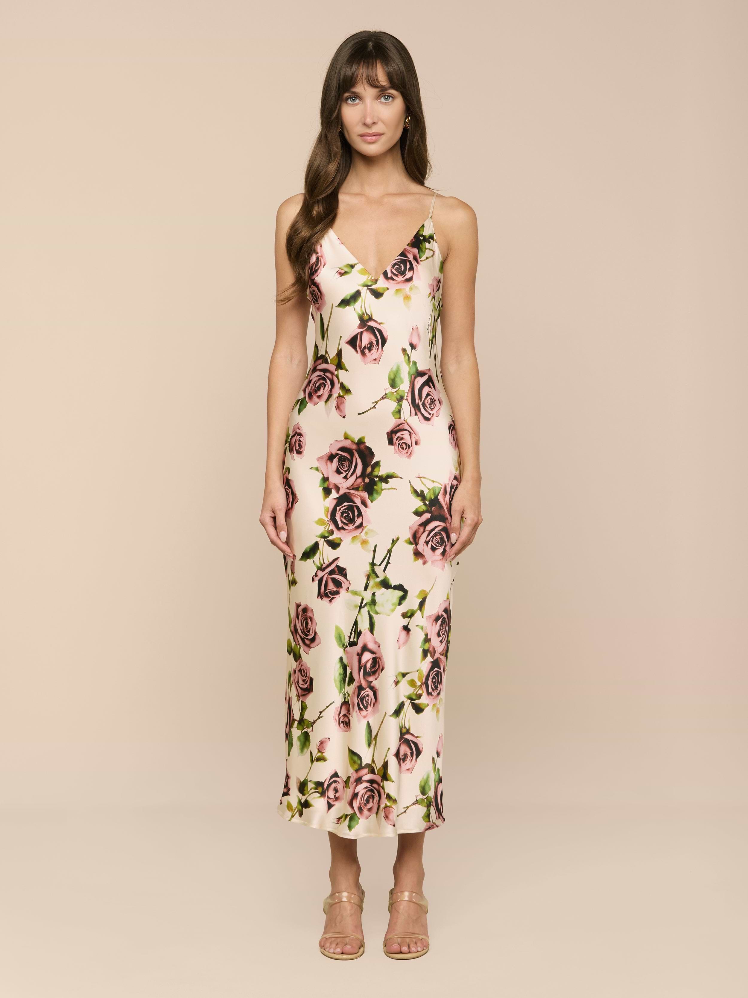 Una mujer con pelo largo castaño y flequillo sobre un fondo beige luce el Seridie Silk Slip Dress, un vestido rosa claro sin mangas con estampado floral de rosas, combinado con unos tacones de tiras en color nude.