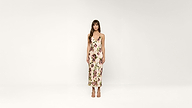 A woman stands against a plain white background, wearing a sleeveless, light-colored midi dress with a floral pattern and light-colored sandals. She has straight brown hair and is looking forward.