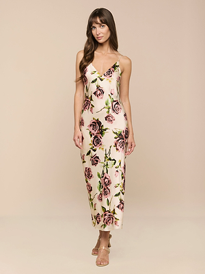A woman with long brown hair wears the Seridie Silk Slip Dress—a bias-cut, ankle-length silk slip featuring a pink and green rose print—paired with strappy sandals, standing against a light beige background.