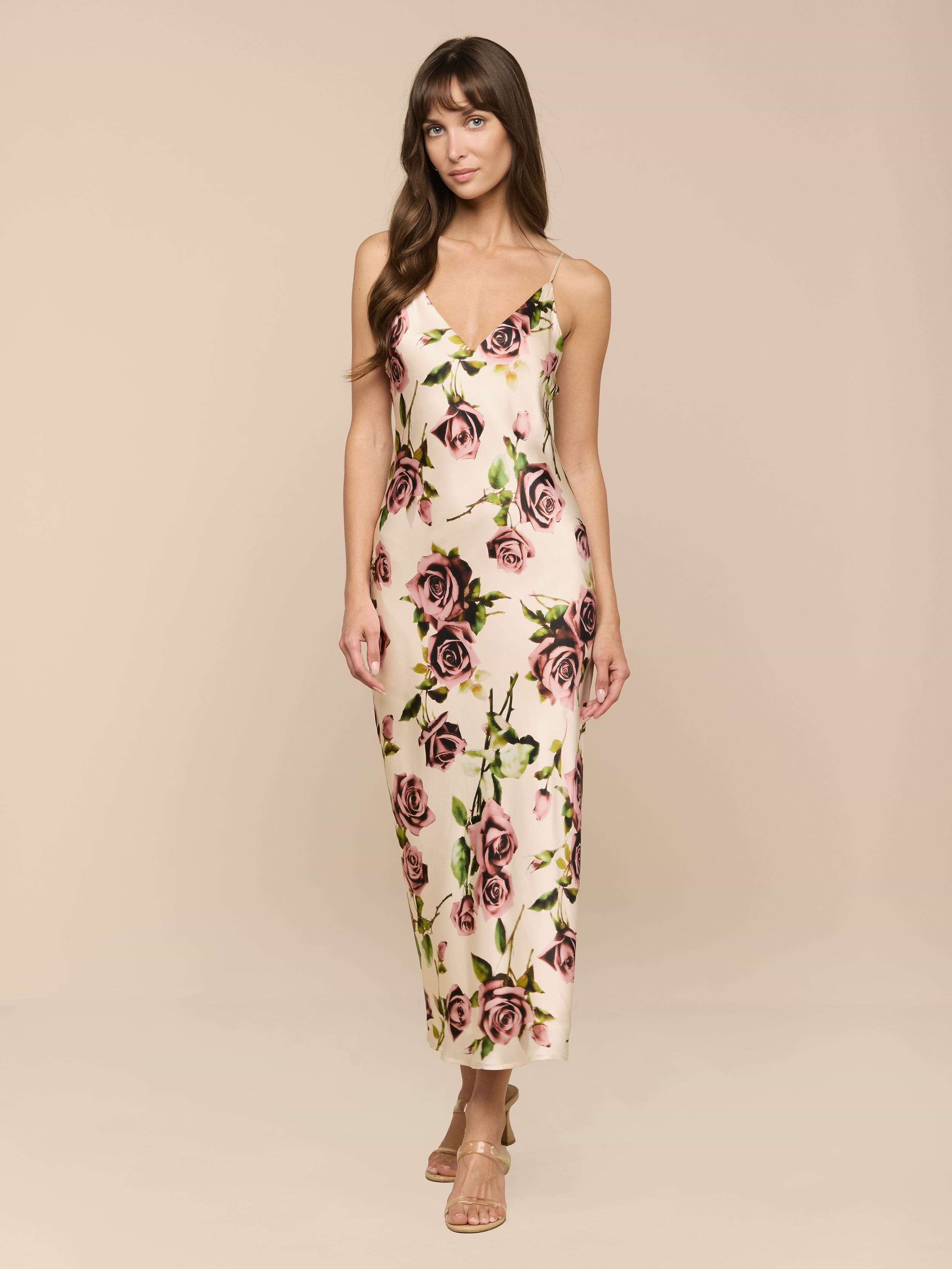 Una mujer con el pelo largo y castaño lleva el vestido Seridie Silk Slip Dress -un slip de seda cortado al bies, largo hasta los tobillos y con un estampado de rosas rosas y verdes- combinado con sandalias de tiras, sobre un fondo beige claro.